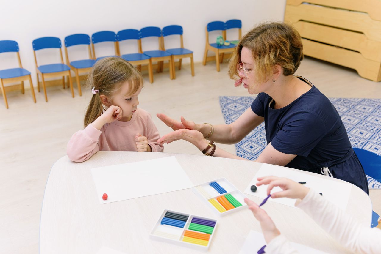 A teacher helps a child with a clay molding activity in a bright kindergarten classroom.